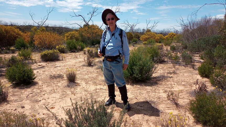 A man in walking gear stands in a dry, shrubby environment.