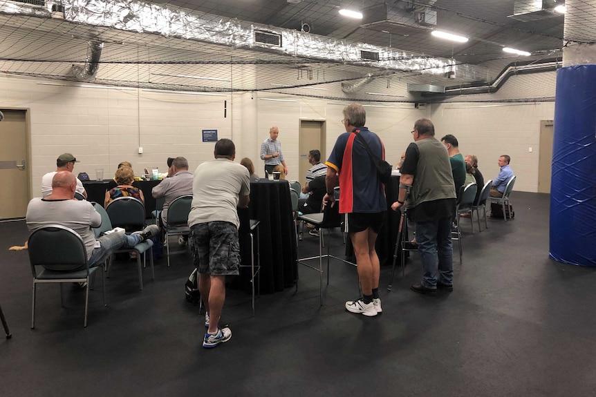 A man stands and addresses seated workers in a stadium changing room.