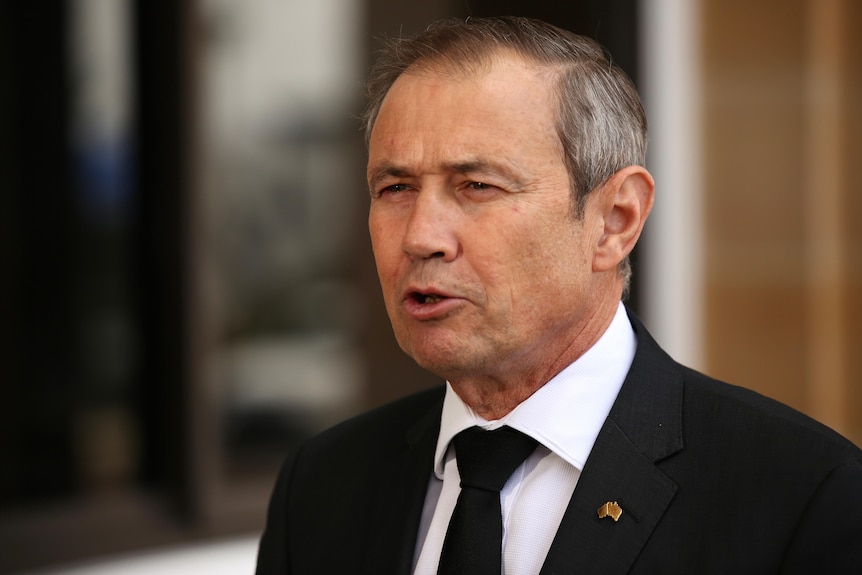 A man in a black suit and tie speaks at a press conference. 