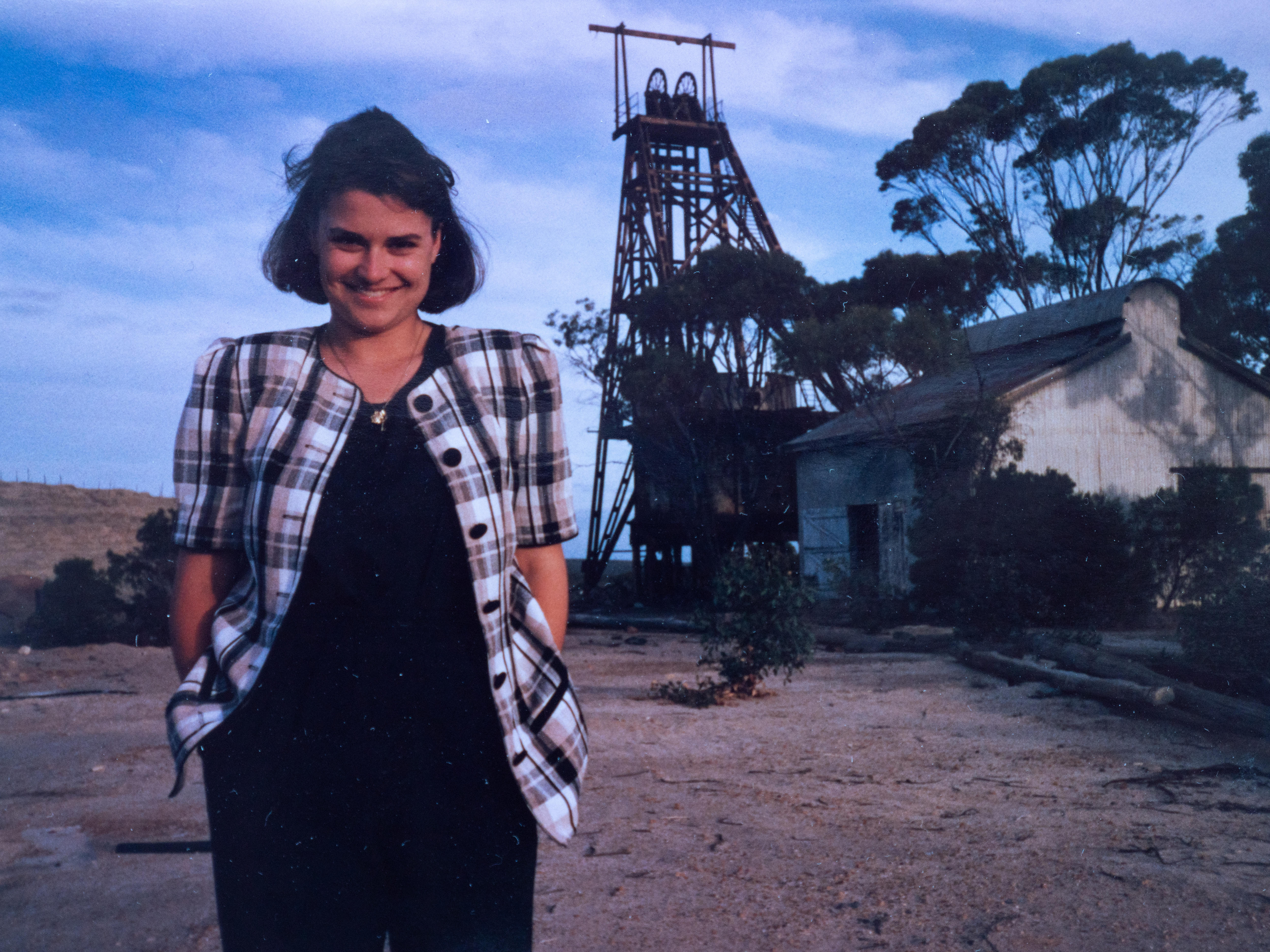 Young woman stands in country area with building and mine shaft in background