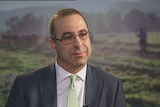 A man, wearing glasses and a suit, sits in front of a picture of a dairy paddock