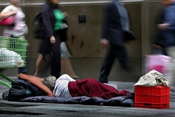 A person lies in a sleeping bag on a footpath as people walk past