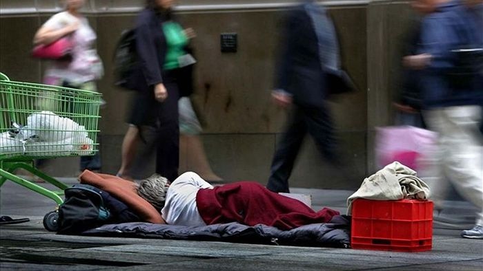 A person sleeps on a footpath next to a shopping trolley. Pedestrians walk past in background.