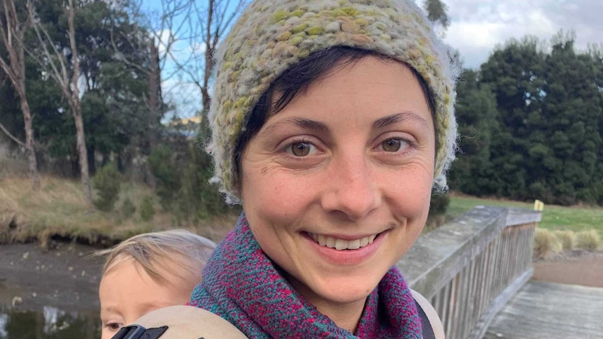 A woman carrying a baby on her back looks at the camera.