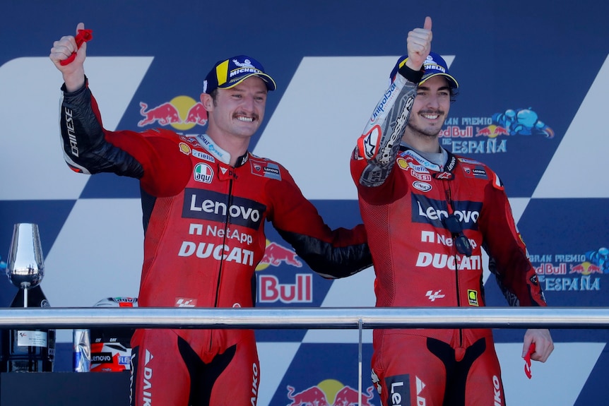 two men in racing uniforms and hats give the thumbs up on a podium