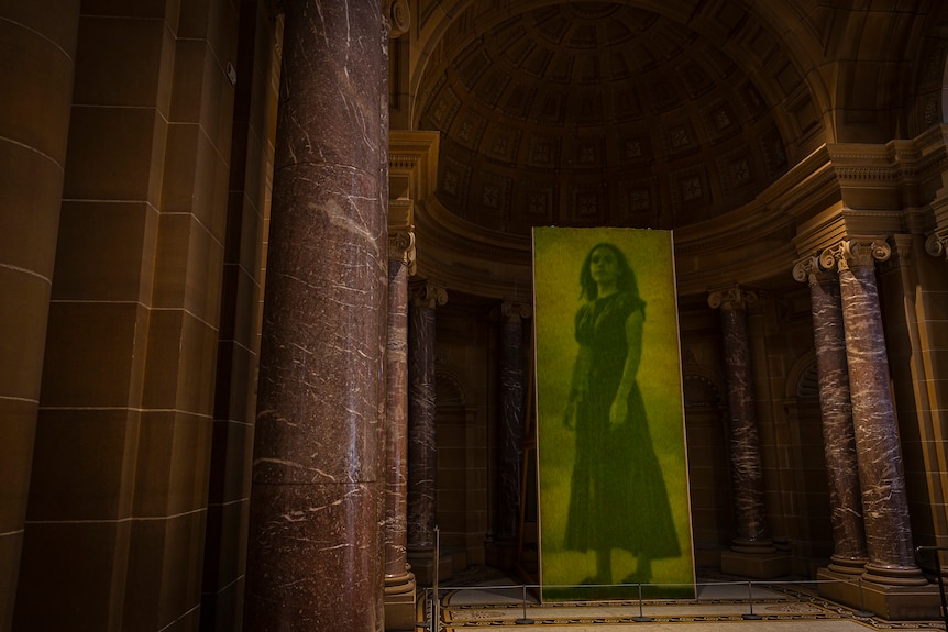 A photographic portrait of a defiant-looking woman printed on grass hanging in a grand-looking foyer