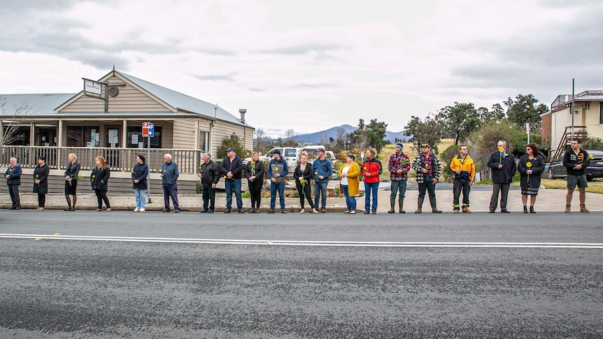 Men and women stand along street of small country town holding yellow flowers, spread evenly with 1 metre between them.