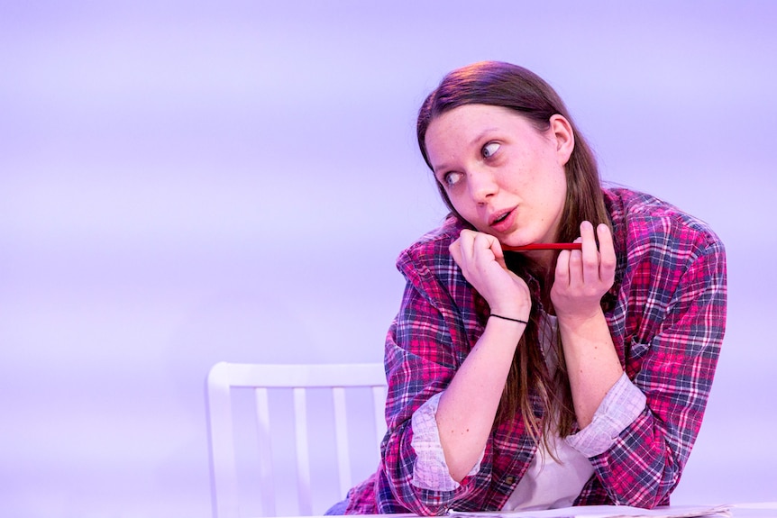 20-ish girl with long hair, wearing flannel button-down over tee shirt, sits at table holding pencil and with paper on table.