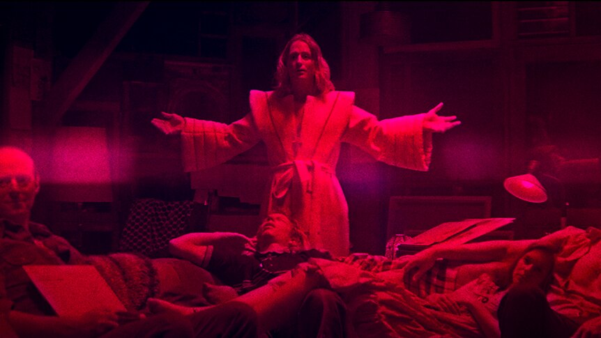 Colour still of Richard Brake, Ned Dennehy, Linus Roache and two others in a red tinted living room in 2018 film Mandy.