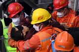 Rescue workers wearing high-vis and hardhats carry a person in their arms.