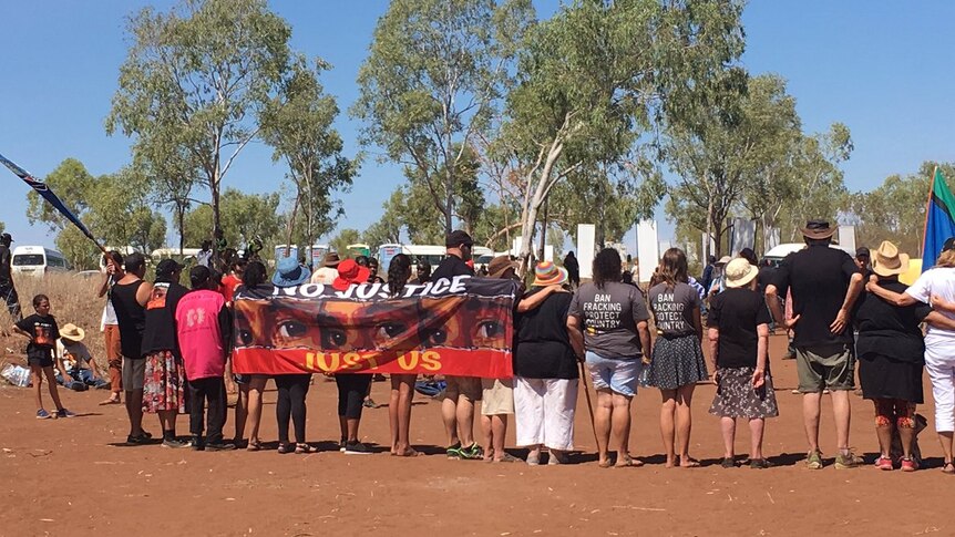 A number of people with their backs turned, holding a banner saying 'no justice, just us'.