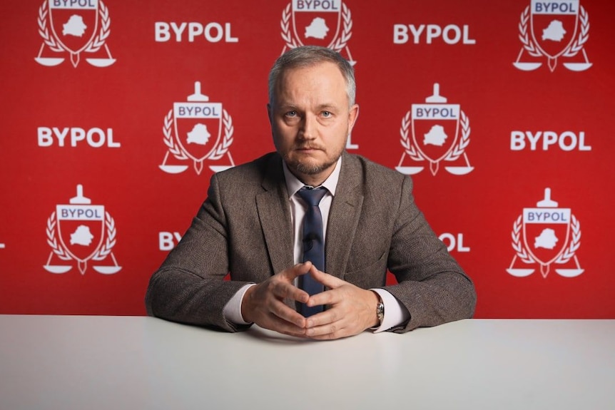 Aliaksandr Azarau sits at a table in a suit with Bypol logos behind him. 