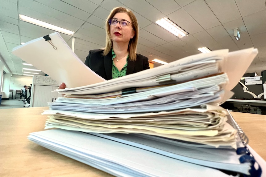 A woman in glasses and a dark jacket and green shirt sits behind a mound of documents in an office.