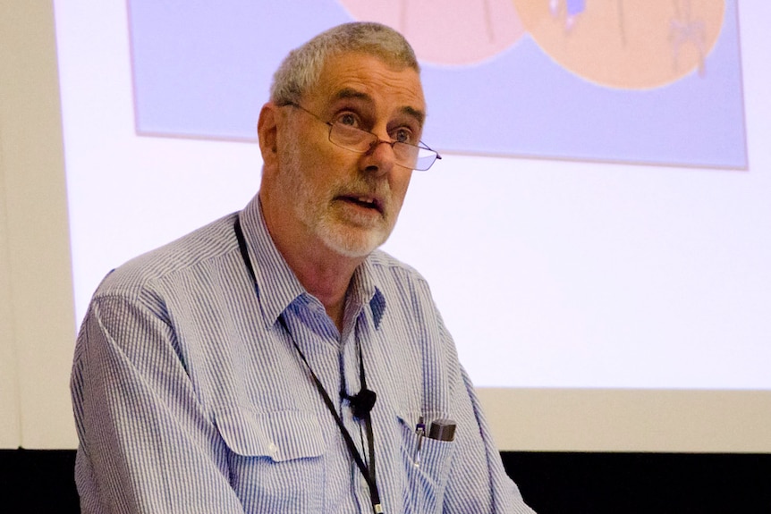 Associate professor Ken Harvey in striped shirt and glasses giving a talk, with a powerpoint image behind him