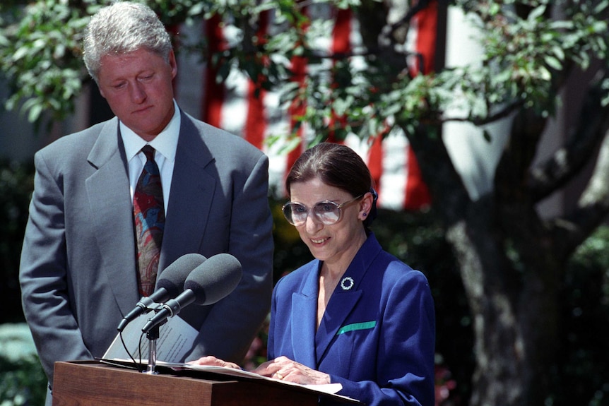 Bill Clinton standing in the Rose Garden, looking down at Ruth Bader Ginsburg as she speaks at a lectern
