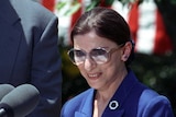 Bill Clinton standing in the Rose Garden, looking down at Ruth Bader Ginsburg as she speaks at a lectern