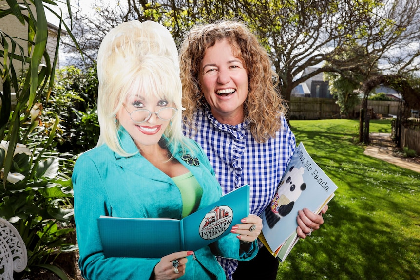 A smiling woman holds a book while standing next to a cardboard cutout of Dolly Parton.