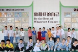 A group of 15 Chinese twins stand in summer clothing in front of a school classroom with Chinese writing