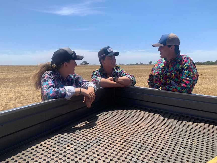 Three teenagers standing a around a ute smiling and talking in a barley paddock with blue sky.
