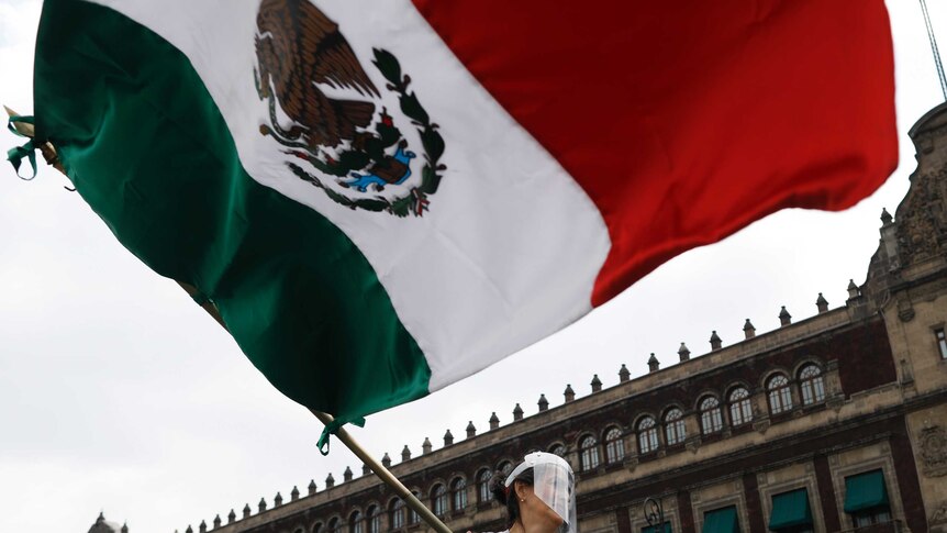 A woman wearing a face shield waves a Mexican flag in front of the National Palace.