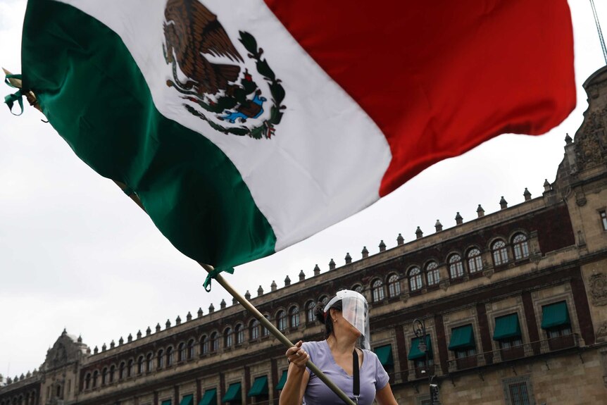 A woman wearing a face shield waves a Mexican flag in front of the National Palace.