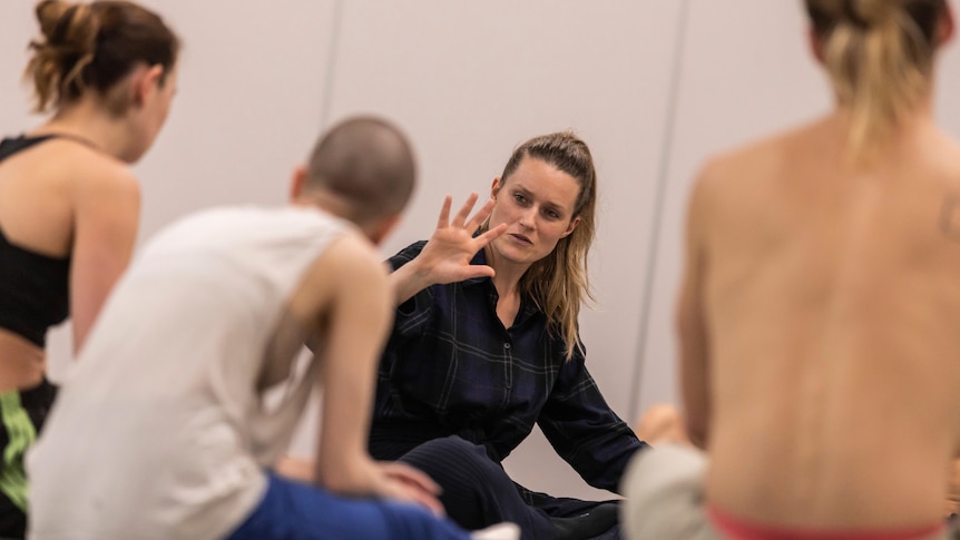 Blonde white woman wearing dark blue flannelette shirt raises hand while speaking to dancers with their backs to the camera.