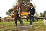 man and woman with spades in a park with an excavator in the background.