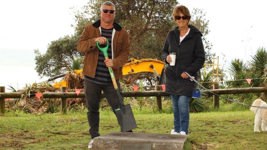 man and woman with spades in a park with an excavator in the background.