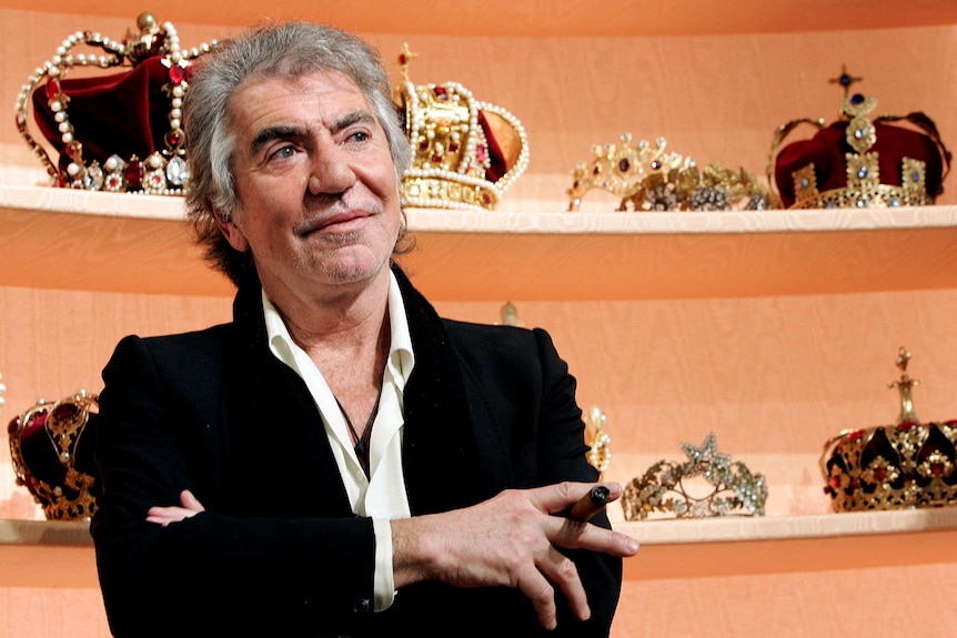 Roberto Cavalli is pictured wearing a black jacket and white shirt while standing with his arms crossed. Behind him are crowns.