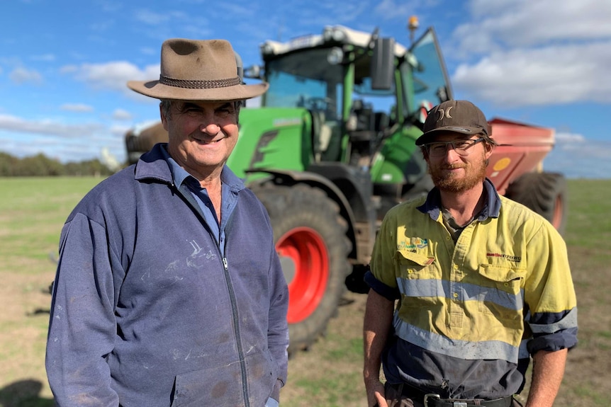 Middle-aged male farmer and young male farm worker standing in front of tractor with fertiliser spreader