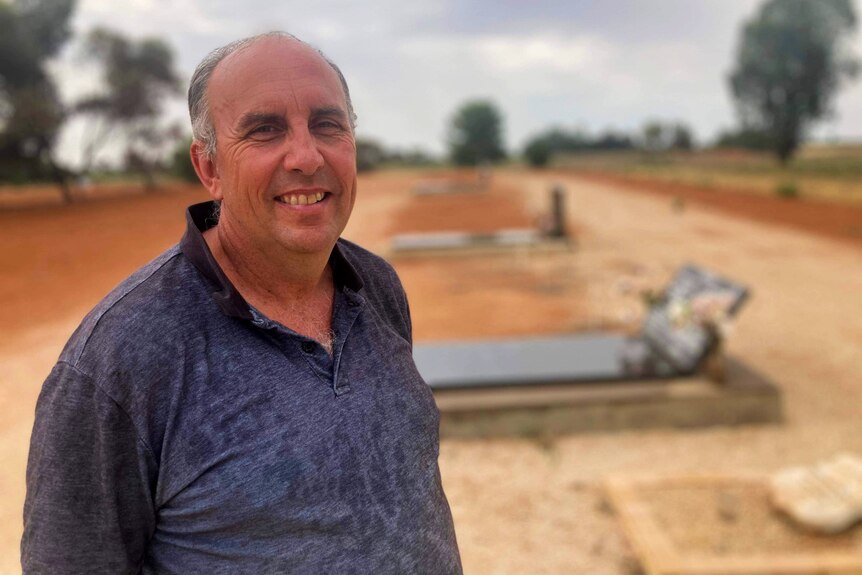 A bald tanned man smiles he wears a dark collared shirt, with gravestones and red dirt behind him