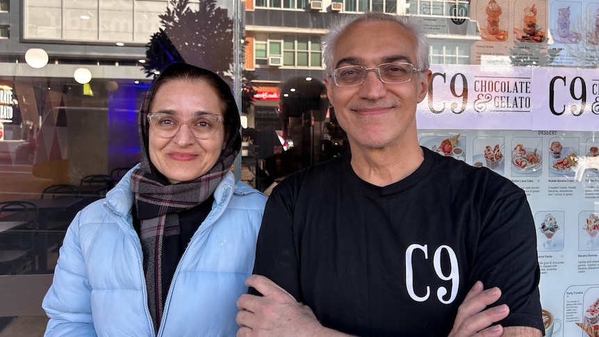 A man and a woman stand smiling outside the window of a gelato store.