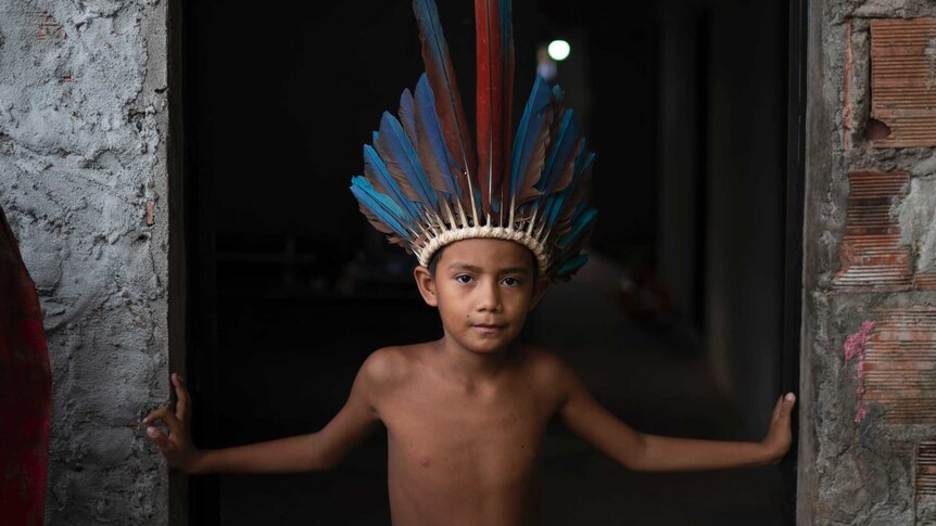 A little indigenous Brazilian boy in a feather head dress looks strong while standing in a doorway
