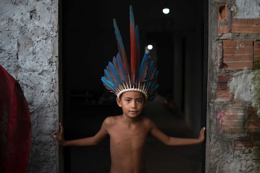 A little indigenous Brazilian boy in a feather head dress looks strong while standing in a doorway