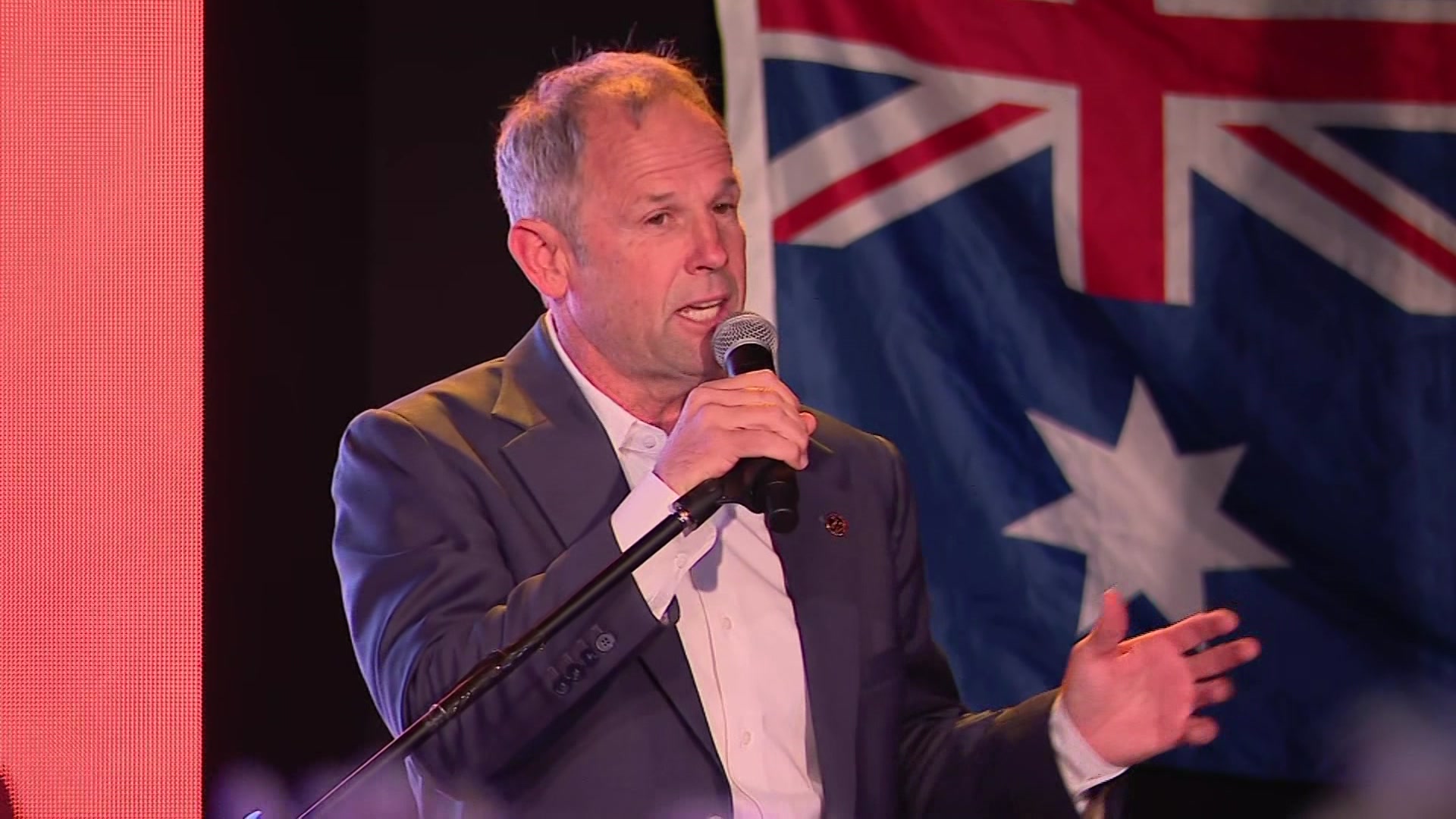 Rod Caddies speaking with a mic in front of an Australian flag.
