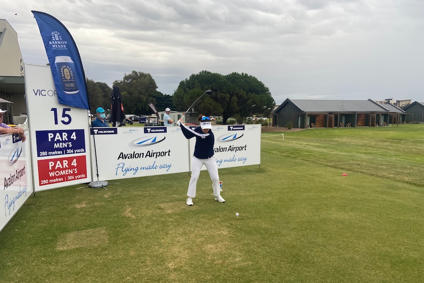 A golf player swings the club backwards at the tee while surrounded by advertising hoardings