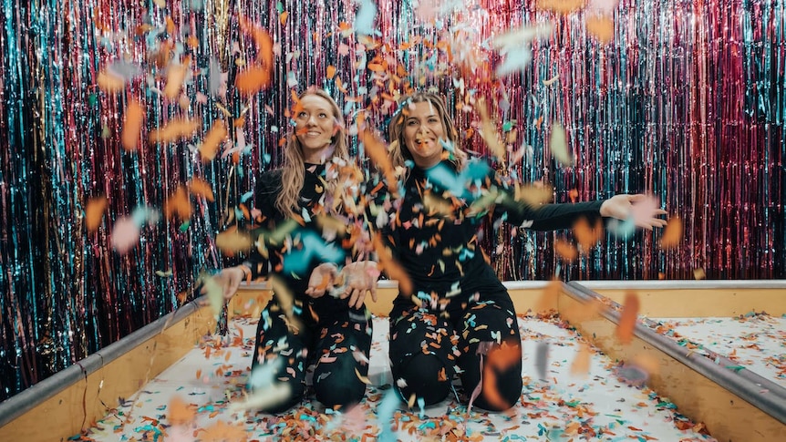 Two women smile as they kneel in a room with streamers and confetti