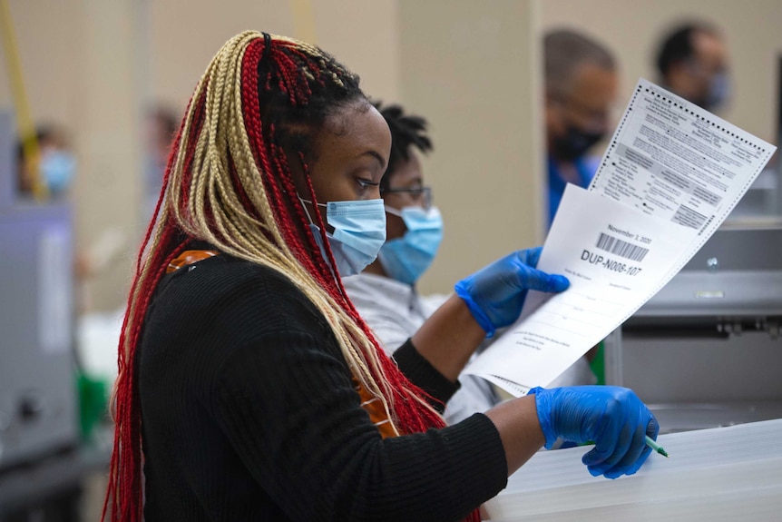 A Black woman with multi-coloured braided hair examines a ballot paper.