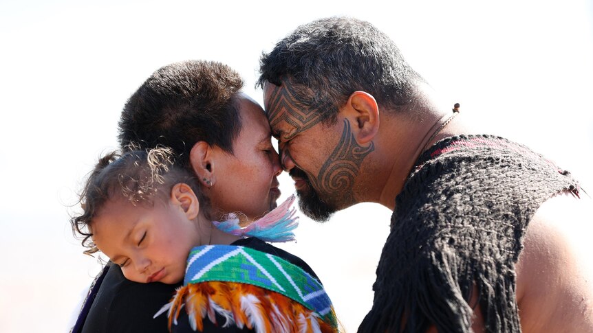 A man and woman embrace with a Māori hongi, nose to nose, while a child sleeps on the woman's shoulder