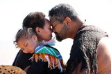 A man and woman embrace with a Māori hongi, nose to nose, while a child sleeps on the woman's shoulder