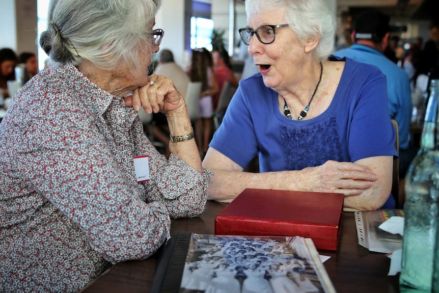 Two older woman sit at a a table indoors talking to one another.
