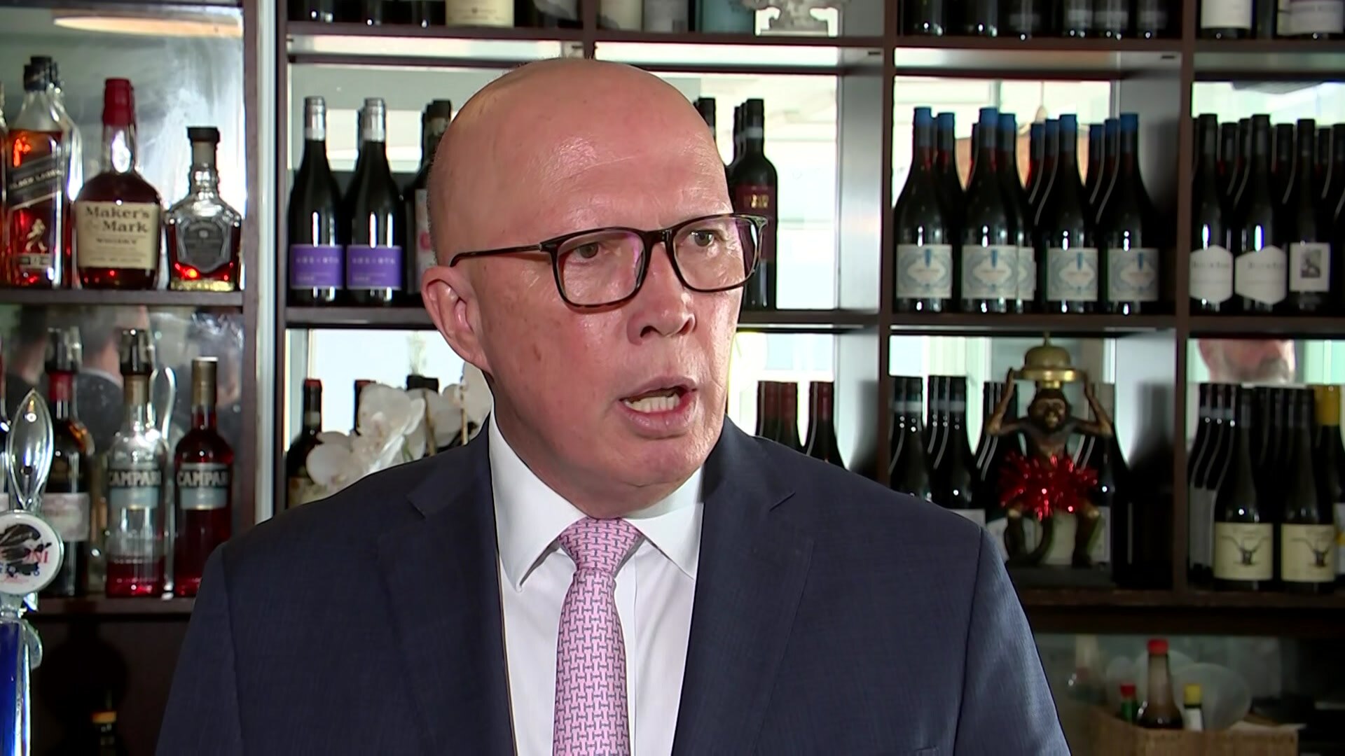 Peter Dutton in a dark suit jacket and pink tie speaking in front of a bar with bottles of alcohol.