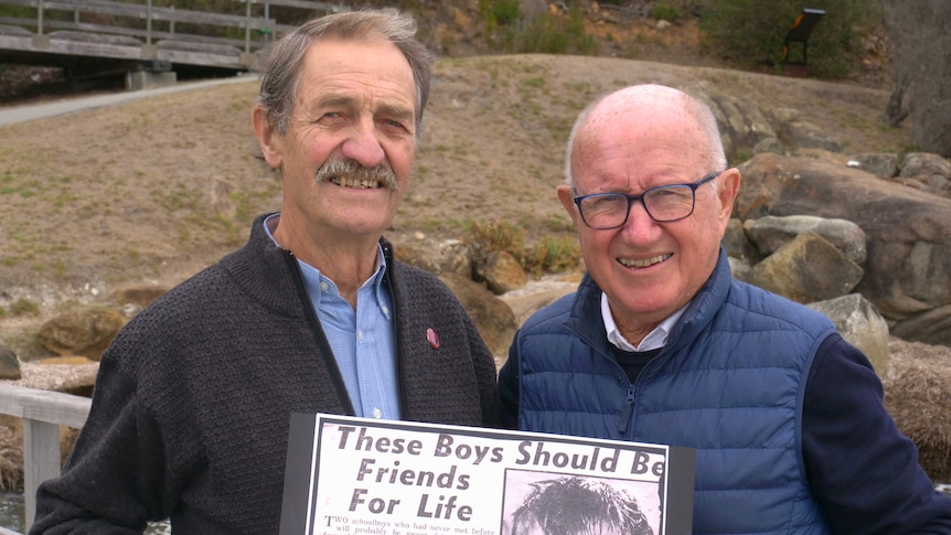 Two men standing together smiling, holding a newspaper article titled "these boys should be friends for life"