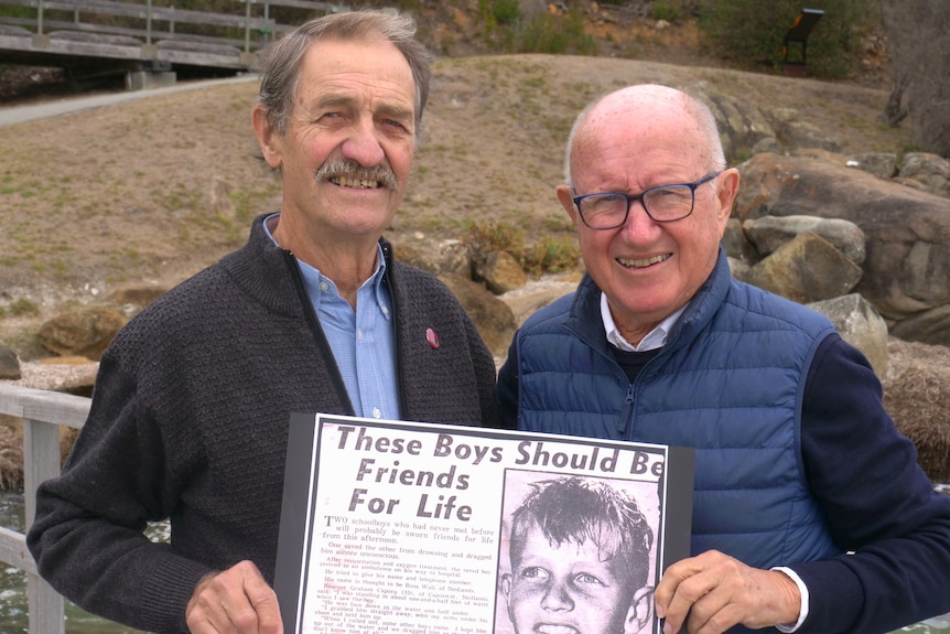 Two men standing together smiling, holding a newspaper article titled "these boys should be friends for life"