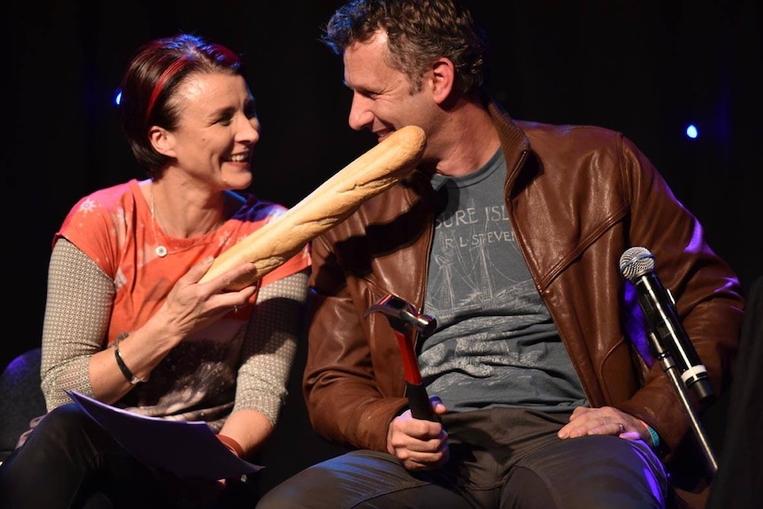 A man and woman smile, holding a baguette and hammer.