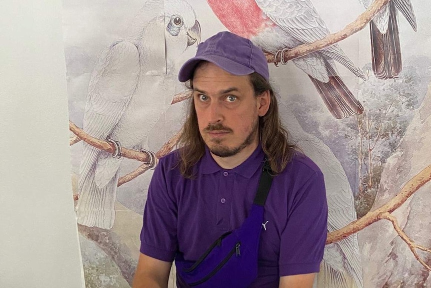 The artist Malcolm Whittaker in purple overalls, tshirt and hat sitting in front a painting of galahs
