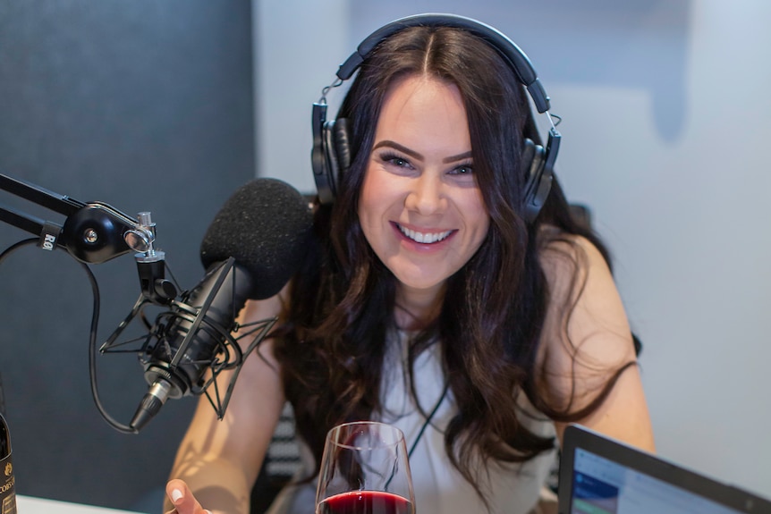 Victoria Devine sits in a sound recording space with a large microphone and a glass of red wine in front of her. 
