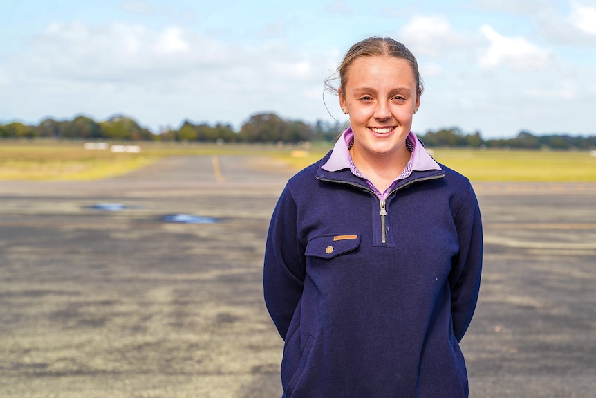 A girl with blond hair wearing a blue jumper stands on a long tarmac smiling.