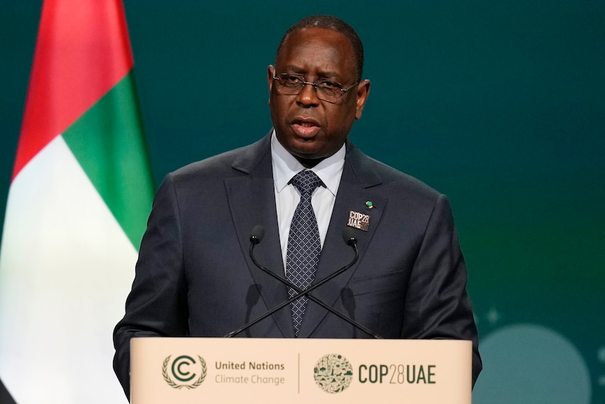 A middle-aged African man in a suit and glasses speaks behind a set of microphones.