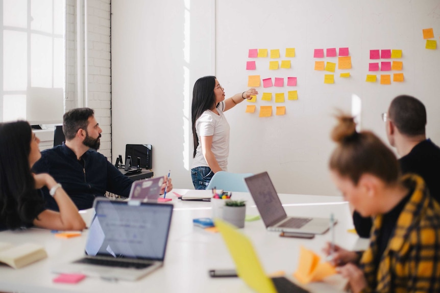 A woman points at sticky notes on a wall while her colleagues sit at a desk.